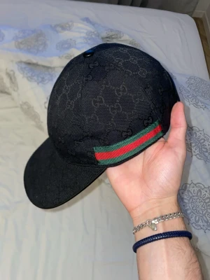 Svart keps från Gucci med GG-mönster - Snygg svart keps från Gucci med klassiskt GG-monogram över hela kepsen. Har en grön och röd rand på sidan och justerbar rem baktill i läder. Perfekt accessoar för att lyfta din outfit med en lyxig touch.