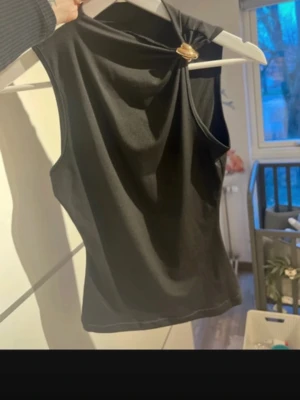 Svart ärmlös topp med gulddetalj - Snygg svart ärmlös topp med unik guldfärgad ringdetalj vid axeln. Toppen har en asymmetrisk design och är tillverkad i ett slätt, stretchigt material som ger en tight passform. Perfekt för att lyfta din outfit med en cool detalj.