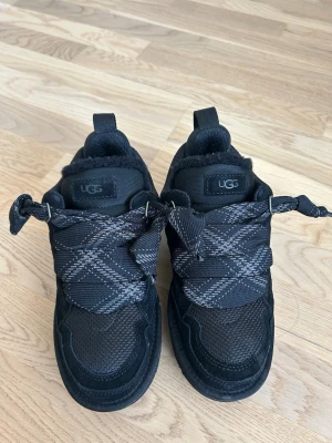 Svarta sneakers från UGG med breda snören - Snygga svarta sneakers från UGG med breda, rutmönstrade snören och detaljer i mocka och mesh. Skorna har en chunky sula och mjukt foder för extra komfort. Använda ca 4ggr så som nya. Kommer med svarta skosnören också och skickas i originalförpackning. Storlek 38