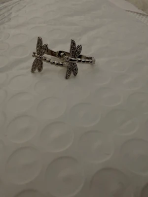 Silverfärgad ring med fjärilar - Unik silverfärgad ring med två fjärilsdekorationer, båda prydda med små gnistrande stenar. Ringen har en pärlad struktur på bandet och en elegant design som sticker ut. Perfekt för dig som gillar detaljer och vill ha något speciellt.