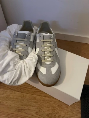 Vita och grå sneakers från Maison Margiela - Säljer ett par stilrena sneakers från Maison Margiela i vitt och ljusgrått. Skorna har ovandel i skinn och mocka, beige snörning och klassisk gummisula. Perfekta till jeans eller dressade byxor för en clean look. Dustbag och original paket med följer, perfekt för sommaren och våren passar bra med allt.