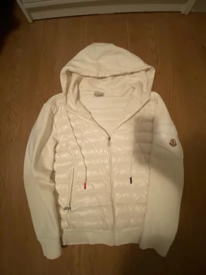 Moncler cardigan  - Snygg vit hoodie från Moncler med quiltad front och ärmficka med logotyp. Dragkedja framtill, två fickor och justerbar huva med snören. Perfekt för dig som vill ha en stilren och exklusiv look. Materialmix av bomull och vadderad nylon.