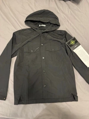 Svart vindjacka från Stone Island - Snygg svart vindjacka från Stone Island med huva och knappstängning framtill. Jackan har två bröstfickor med lock och ikonisk Stone Island-patch på ärmen. Tillverkad i lätt och slitstarkt material med multilager fusionsteknologi.