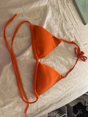Orange bikini med knyt i sidorna - Snygg orange bikini med trekantsöverdel och knyt i nacken och ryggen. Underdelen har knyt i sidorna för justerbar passform. Båda delarna är ribbade och har en fräsch, ungdomlig look – perfekt för stranden eller poolen.