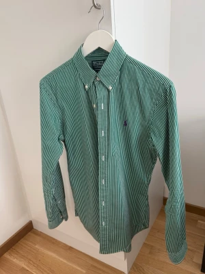 Grönrandig skjorta från Polo Ralph Lauren - Klassisk grön och vit randig skjorta från Polo Ralph Lauren med button-down krage och broderad logga på bröstet. Skjortan har lång ärm och knäppning framtill. Perfekt för en stilren look. I storlek S