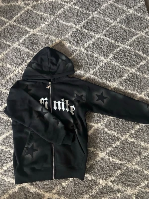 Svart hoodie med stjärnor och tryck - Säljer en svart hoodie med dragkedja från Infinite. Hoodien har stora vita bokstäver på bröstet och flera svarta stjärnor tryckta både fram och bak. Utrustad med huva och två fickor framtill. Perfekt för dig som gillar streetwear och coola detaljer.