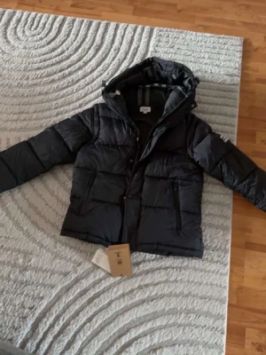 Svart pufferjacka från Burberry - Snygg svart pufferjacka från Burberry London England med stor huva och dragkedja framtill. Jackan har två stora fickor med dragkedja och en tydlig Burberry-logga på ärmen. Perfekt för kalla vinterdagar och riktigt trendig look.