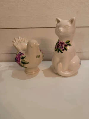 Prydnadsfigur katt och fågel keramik - Två dekorativa prydnadsfigurer i keramik, en sittande katt och en fågel. Båda är vita med handmålade blommor i lila och grönt på kroppen. Perfekta som inredningsdetaljer för att ge rummet en personlig touch.