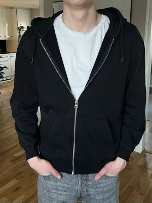 Svart zip hoodie med huva - Klassisk svart hoodie med dragkedja framtill och huva med dragsko. Hoodien har två stora fickor på magen och ribbade muddar vid ärmslut och nederkant. Perfekt för en avslappnad och stilren look. Materialet känns mjukt och bekvämt, troligen bomullsblandning. (Storlek small, stor i storlek)