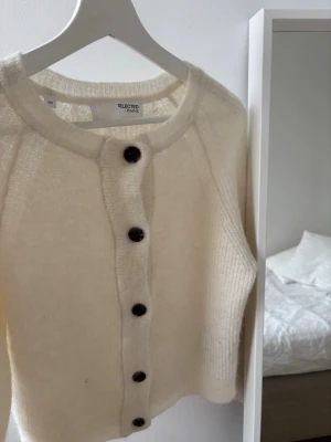 Vit kofta från Selected Femme - Mysig vit kofta från Selected Femme med rund halsringning och svarta knappar framtill. Koftan är stickad i ett fluffigt material och har långa ärmar. Perfekt för lager på lager och en clean look.