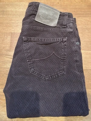 Bruna jeans från Jacob Cohen - Snygga bruna jeans från Jacob Cohen med diskret mönster och klassisk femficksmodell. Jeansen har silverfärgade knappar och patch med logga bak i midjan. Perfekta för dig som vill ha något unikt men ändå stilrent.
