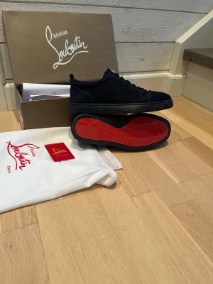 Mörkblåa Christian Louboutin sneakers - Snygga mörkblå sneakers från Christian Louboutin med ikonisk röd sula och diskret logga. Skorna har en klassisk låg profil och är tillverkade i mocka med svarta snören. Perfekta för dig som vill ha en exklusiv och stilren look. Storlek 42. Har en lite skavank på ena sulan.