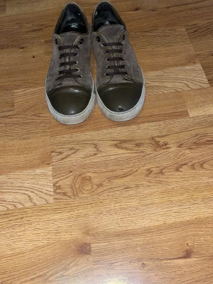 Bruna lanvins mocka  - Säljer ett par bruna sneakers i mocka med rund tå och tåhätta i mörkgrönt gummi. Skorna har vita sulor och bruna snören. Snygga detaljer med metallöljetter och kontrastsömmar. Passar perfekt till jeans eller chinos.