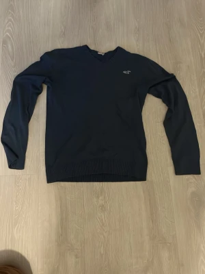 Mörkblå V-ringad tröja Hollister - Mörkblå V-ringad tröja från Hollister med diskret logga broderad på bröstet. Tröjan har långa ärmar och ribbade muddar vid ärmslut och nederkant. Perfekt för lager-på-lager och enkel att matcha med jeans eller chinos.