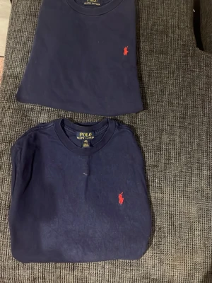Mörkblå t-shirt från Polo Ralph Lauren - Två mörkblå t-shirts från Polo Ralph Lauren med klassisk rund hals och röd broderad logga på bröstet. T-shirten har en enkel och stilren design, perfekt för en avslappnad look. Ena har ett hål i baksidan ej synbart. Båda är 12 års åldern m storlek 400 för båda 