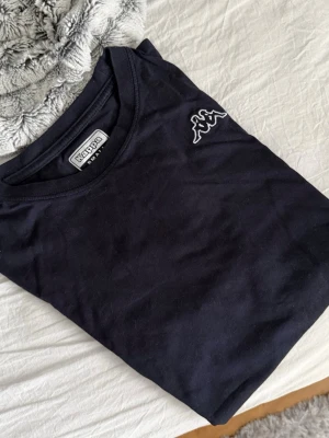 Svart t-shirt från Kappa - Svart t-shirt från Kappa med klassisk rund halsringning och vit broderad logga på bröstet. Tillverkad i mjuk bomull. Enkel och stilren design som passar till det mesta.