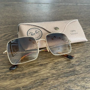 Ray-Ban fyrkantiga solglasögon guld - Ray-Ban (square 1971) solglasögon  med fyrkantiga glas och tunn guldfärgad metallbåge. Glasen är tonade i brunt. Kommer med original Ray-Ban-fodral i beige. 
