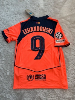Barcelona Lewandowski matchtröja orange - Snygg orange FC Barcelona matchtröja med Lewandowski och nummer 9 på ryggen. Tröjan har mörkblå detaljer, klubbmärke och Spotify-logga på bröstet samt Champions League-märke på ärmen. Tillverkad i lätt och ventilerande material, perfekt för fotboll.