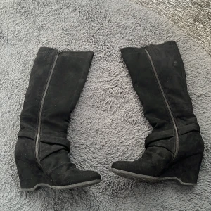 Svarta kilklackstövlar med dragkedja - Säljer ett par svarta stövlar med kilklack och dragkedja på sidan. Stövlarna är i mockaimitation och har en snygg, slank form som går upp till knäna. Perfekta för att lyfta vilken outfit som helst.