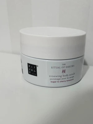 Rituals Sakura Body Scrub - En lyxig kroppsskrubb från Rituals, The Ritual of Sakura, med socker och körsbärsblom. Kommer i en vit rund burk med skruvlock. Skrubben exfolierar huden och lämnar den mjuk och väldoftande.