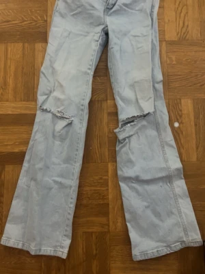 Ljusblå bootcut jeans - Säljer ett par ljusblå bootcut jeans med slitningar och hål på båda benen. Jeansen har klassisk femficksdesign och gylf med knapp. Perfekta för en avslappnad och trendig look.