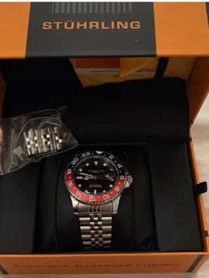 Stührling klocka med blå och röd bezel - Snygg Stührling klocka med silverfärgad länkarmband i metall och en cool blå och röd bezel. Svart urtavla med tydliga vita markeringar och datumfönster. Kommer i originalbox och extra länkbitar medföljer.Fint skick och använd med försiktighet