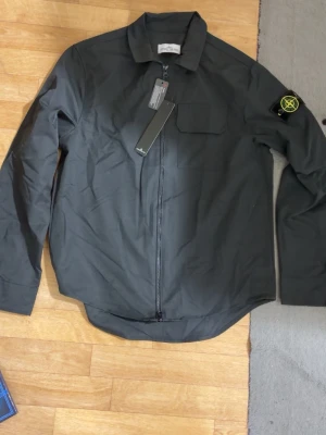 Svart overshirt från Stone Island - Snygg svart overshirt från Stone Island med dragkedja framtill, klassisk krage och bröstficka. Ikonisk logotyp-patch på vänster ärm. Tillverkad i ett lätt och slitstarkt material som passar perfekt till lager-på-lager-stil.