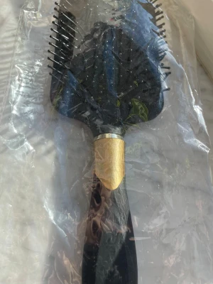 Svart paddle hårborste med gulddetalj - Stor paddle hårborste i svart plast med guldfärgad detalj vid handtaget. Borsten har bred, rektangulär form och flexibla piggar med runda toppar som är skonsamma mot hårbotten. Perfekt för att reda ut långt eller tjockt hår utan att slita.