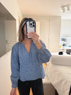Blå broderad blus med lång ärm - Snygg blå blus med broderade hålmönster och v-ringning. Blusen har långa ärmar med resår vid handlederna och en avslappnad passform. Perfekt för dig som gillar en trendig och bekväm stil.