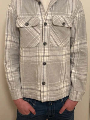 Grå rutig overshirt från Jack & Jones - Säljer en stilren grå overshirt från Jack & Jones med vitt rutmönster. Jackan har två bröstfickor med knappar och klassisk krage. Perfekt att slänga över en t-shirt för en avslappnad look. Tillverkad i mjuk bomull och stängs med knappar framtill. Storlek XS passar på längden 164-170