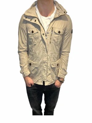 Beige field jacket från Peak Performance - Stilren beige field jacket från Peak Performance med flera fickor framtill, bruna knappar och dragkedja. Jackan har hög krage och patch på ärmen. Perfekt för dig som vill ha en snygg och funktionell jacka till vardags.