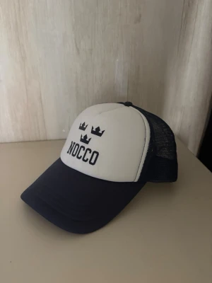 NOCCO blåvit truckerkeps - Snygg blåvit truckerkeps med NOCCO tryck med mörkblå mesh på sidorna och bak, samt böjd skärm. Framsidan har NOCCO-logga och tre kronor tryckta i mörkblå . Perfekt för en sportig och avslappnad stil. Endast använd EN gång