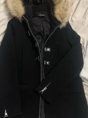 Svart kappa med fuskpäls från Otherwear - Stilren svart kappa med huva och fuskpäls från Otherwear. Kappan har dragkedja, dekorativa spännen framtill och fickor på sidorna. Perfekt för kyliga dagar och ger en snygg siluett. Fodrad insida för extra värme.