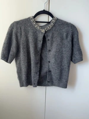 Grå kofta med pärldetaljer H&M - Kortärmad grå kofta från H&M med rund halsringning dekorerad med pärlor och glittriga stenar. Koftan har knäppning framtill och är stickad i ett mjukt material, perfekt för lager-på-lager-stil.