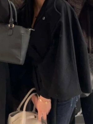 Svart cape-rock med knytband - Säljer en svart poncho med knytband i midjan och vida ärmar. Rocken har hög krage och en stilren, minimalistisk design som passar perfekt till jeans. Endast använd en gång och säljs för jag vill köpa en annan! 