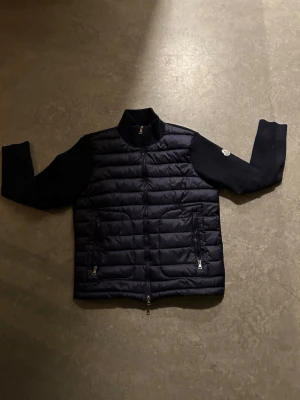 Mörkblå dunväst från Moncler - Snygg mörkblå dunväst från Moncler med quiltad front och två dragkedjeförsedda fickor. Västen har stickade ärmar och hög krage, samt en Moncler-logga på ena ärmen. Perfekt för lager-på-lager och streetstyle.