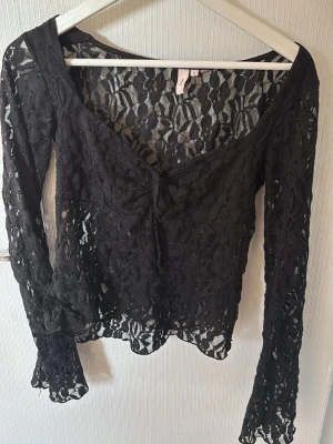 Svart spetsblus  - Säljer en svart långärmad blus från Nelly i genombruten spets med v-ringning och knyt framtill. Blusen är figurnära och har utsvängda ärmar, perfekt för en trendig look.