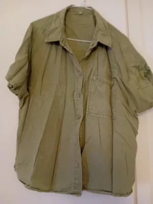 Olivgrön skjorta med kort ärm - Olivgrön skjorta med kort ärm och klassisk krage. Skjortan har en bröstficka och knäppning framtill. Perfekt för en avslappnad stil och enkel att matcha med jeans eller shorts.