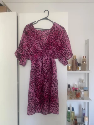 Rosa leopardmönstrad klänning - Snygg rosa klänning med leopardmönster och vida korta ärmar. Klänningen har en markerad midja och är i en kort modell. Perfekt för dig som vill sticka ut med ett djurmönster och en färgstark look.
