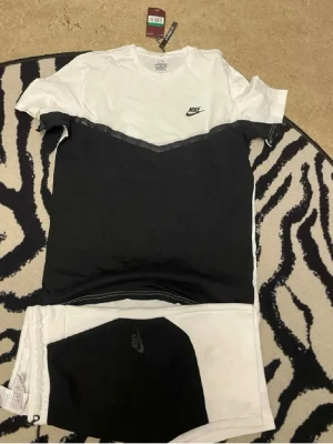 Nike svartvitt set med t-shirt & shorts - Snyggt set från Nike med en svart och vit t-shirt med V-formad färgövergång och matchande shorts. T-shirten har rund hals och korta ärmar, Nike-logga på bröstet. Shortsen är vita med svart detalj och Nike-logga. Perfekt för en sportig look.