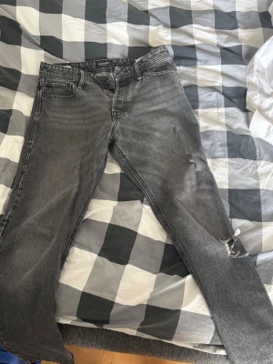 Jeans Jack and Jones  - Säljer ett par grå svarta jeans från Jack and Jones, modell Loose/Chris. Jeansen har en snygg sliten look och med ett hål på ena knät som blivit efter användning. Utöver det är dom i grymt skick. 