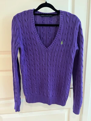 Lila kabelstickad tröja från Ralph Lauren - Lila kabelstickad tröja från Ralph Lauren med V-ringning och lång ärm. Tröjan har en liten grön broderad logga på bröstet och ribbade muddar vid ärmslut och nederkant. Perfekt för dig som gillar klassisk och färgstark stil.