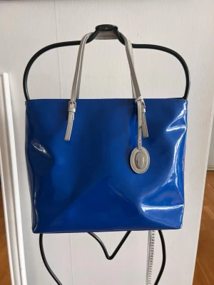 Blå shoppingväska med silverhandtag - Snygg shoppingväska i blankt blått material med stilrena silverfärgade handtag. Väskan har en klassisk fyrkantig form och ett dekorativt hänge framtill. Perfekt för dig som vill sticka ut med en färgstark accessoar.