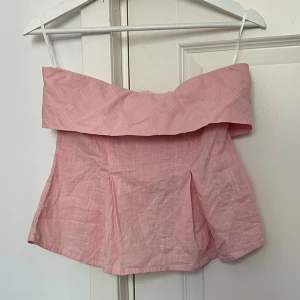 Ljusrosa axelbandslös topp - Söt ljusrosa axelbandslös topp med bred volang upptill och lätt utsvängd passform. Tillverkad i ett tunt, luftigt bomullstyg som passar perfekt till varma dagar. Toppen har en bandeau-liknande form och stängs med dragkedja bak. Lite för stor för mig. 