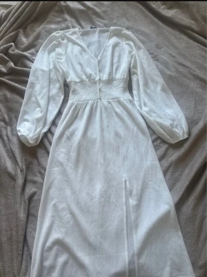 Vit långklänning med v-ringning - Superfin tunn vit långklänning med v-ringad hals och långa ärmar. Klänningen har markerad midja med smock och en slits framtill i lätt strukturerad tyg. Perfekt för sommarens alla tillfällen! Något genomskinligt