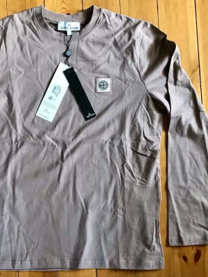 Beige långärmad topp från Stone Island - Snygg beige långärmad topp från Stone Island med rund halsringning och ikonisk logotyp på bröstet. Tillverkad i mjuk bomull som känns skön mot huden. Perfekt för dig som gillar stilrena och enkla plagg med exklusiv känsla.