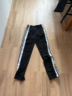 Svarta Adidas mjukisbyxor med ränder - Svarta mjukisbyxor från Adidas med klassiska vita ränder längs sidorna och broderad logga. Byxorna har elastisk midja och är tillverkade i mjukt material som passar perfekt för en avslappnad stil. Snygg detalj med tryckknappar längs benen. Är lite smutsiga längst ner