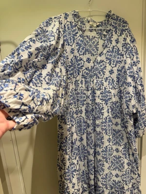 Nyskick! Blåvit långklänning från Indiska - Super fin!! Luftig långklänning från Indiska i vitt med blått blommigt mönster. Klänningen har v-ringning, volangdetaljer vid halsen och långa puffärmar med resår vid ärmslut. Perfekt för en somrig look! Från indiska i stl XL. Möjligen är den lite skrynklig då den legat bland bortsorterade kläderna. Annars aldrig använd.