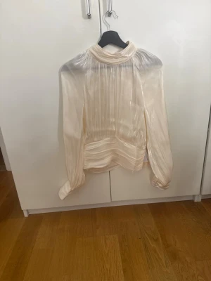 Krämvit plisserad blus med hög krage - Elegant krämvit blus med plisserade detaljer. Blusen har långa ärmar och breda manschetter, samt ett glansigt material som ger en lyxig känsla. Perfekt för dig som gillar en chic och stilren look.