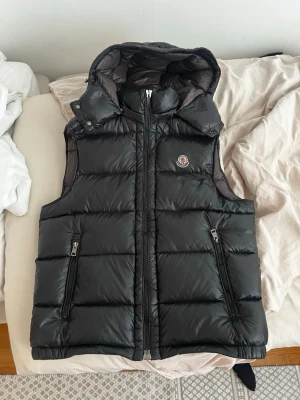Svart dunväst från Moncler med huva - Snygg svart dunväst från Moncler med glansig finish, två dragkedjefickor och avtagbar huva. Klassisk Moncler-logga på bröstet och quiltad design. Perfekt för vilken typ av väder dragkedja funkar som den ska men själva handtagen är borta men det är inget man märker 